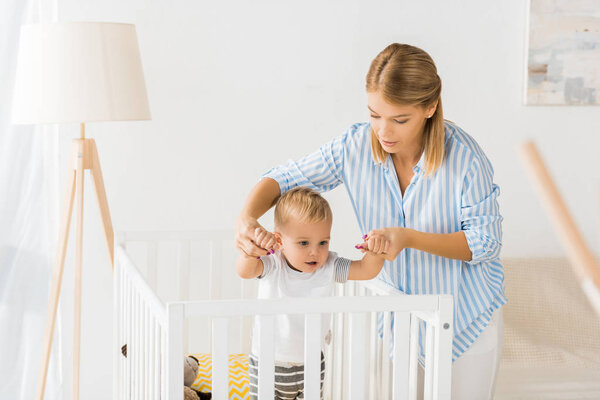 mom holding hands of son in crib in nursery room