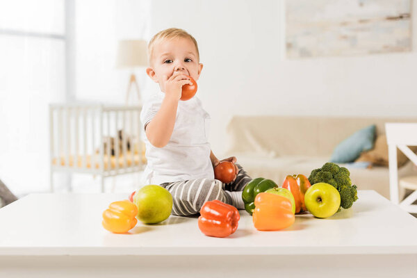 cute toddler eating and sitting on table surrounded by fruit and vegetables