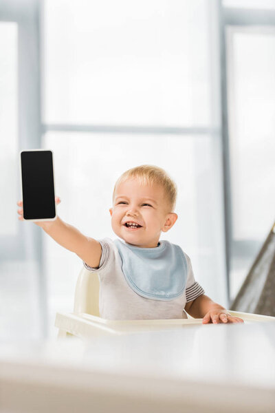 cute smiling toddler holding smartphone in hand and sitting in baby chair