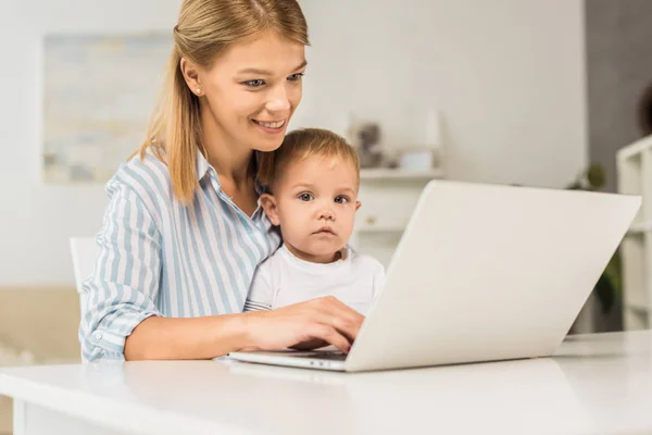 母亲坐在办公桌与可爱的幼儿 而使用笔记本电脑 — 图库照片
