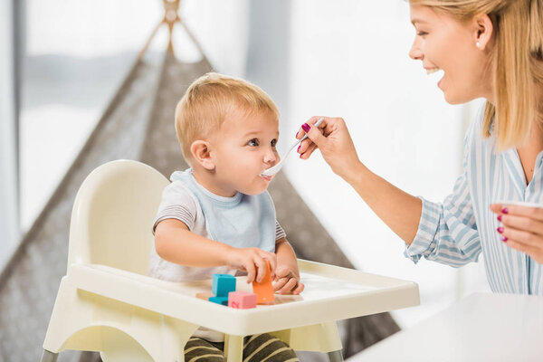happy mom feeding son in highchair with baby food 