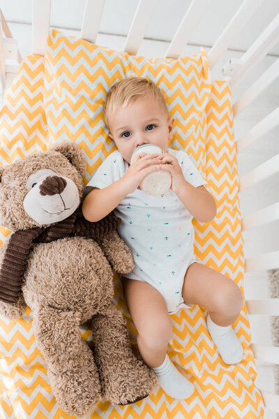 adorable toddler in crib with fluffy teddy bear toy drinking out of baby bottle in nursery room