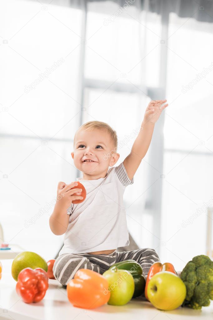 cute toddler raising hand and sitting on table surrounded by fruit and vegetables 