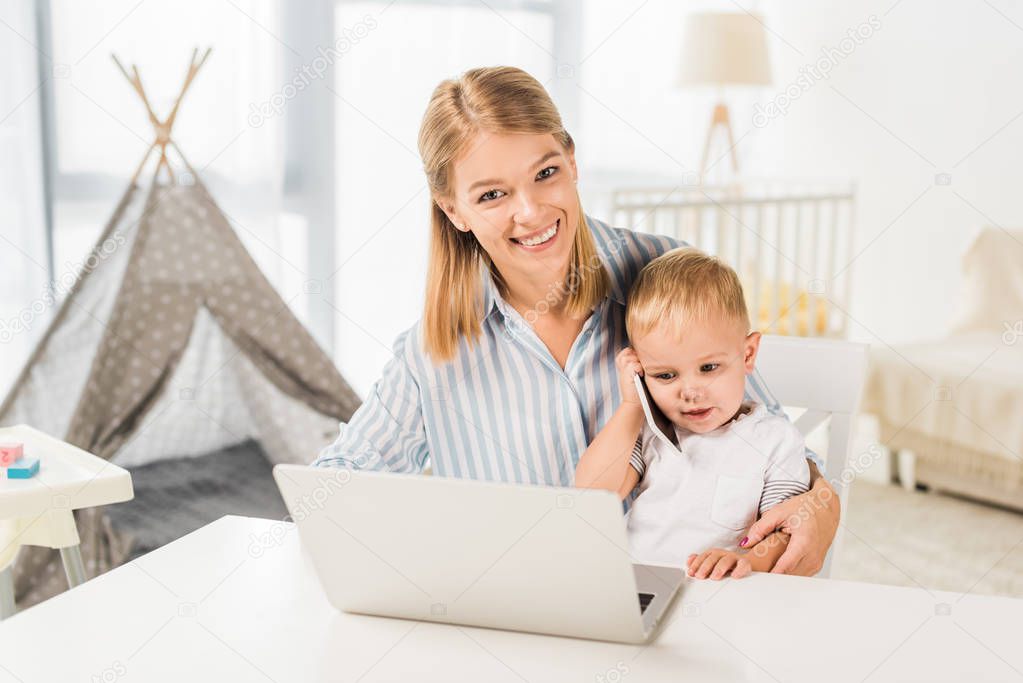 cute toddler holding smaptphone with mother sitting at desk while using laptop
