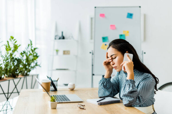 stressed asian businesswoman sitting at computer desk and talking on smartphone in office