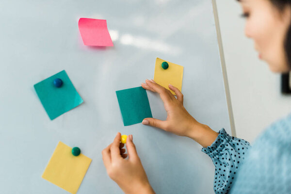cropped view of woman putting colorful sticky notes on white board in office