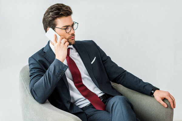 handsome businessman in formal wear sitting on armchair and talking on smartphone in office