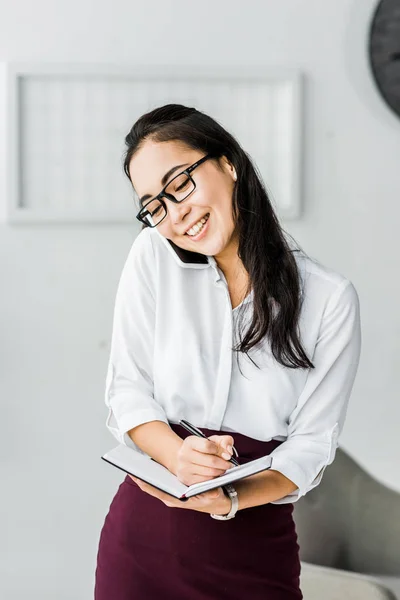 Smiling Asian Businesswoman Talking Smartphone Writing Notebook Office — Stock Photo, Image