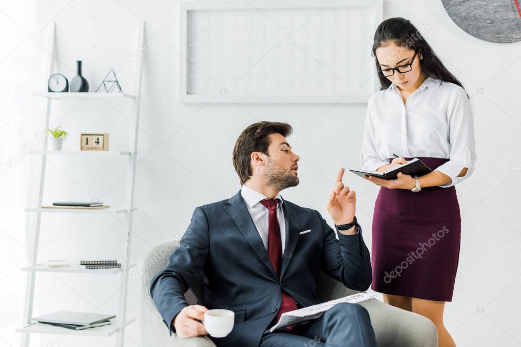 asian businesswoman writing in notebook while businessman sitting and having coffee break in office
