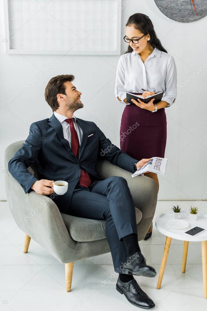 asian businesswoman writing in notebook while businessman sitting on armchair with newspaper and coffee in office