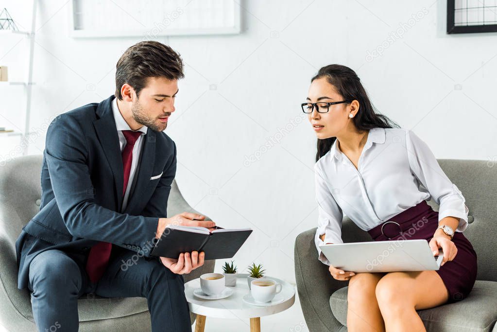 multiethnic businesspeople with notebook and laptop talking while sitting in waiting hall