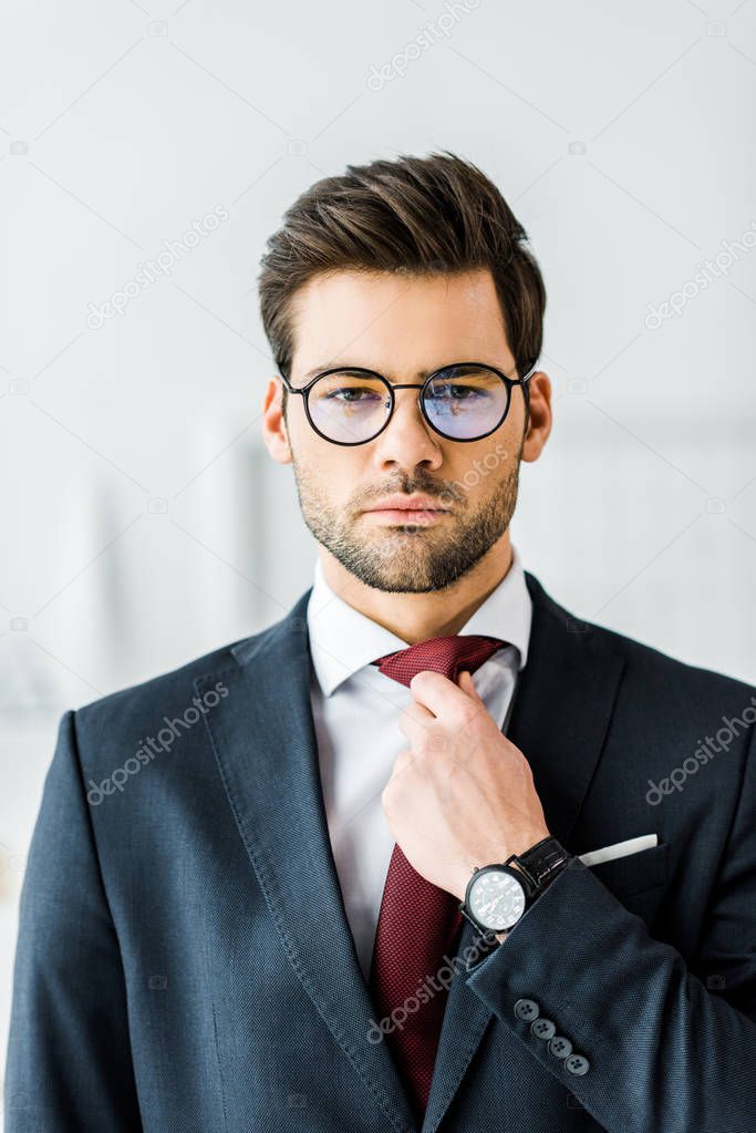 handsome businessman in formal wear and glasses adjusting tie while looking at camera in office