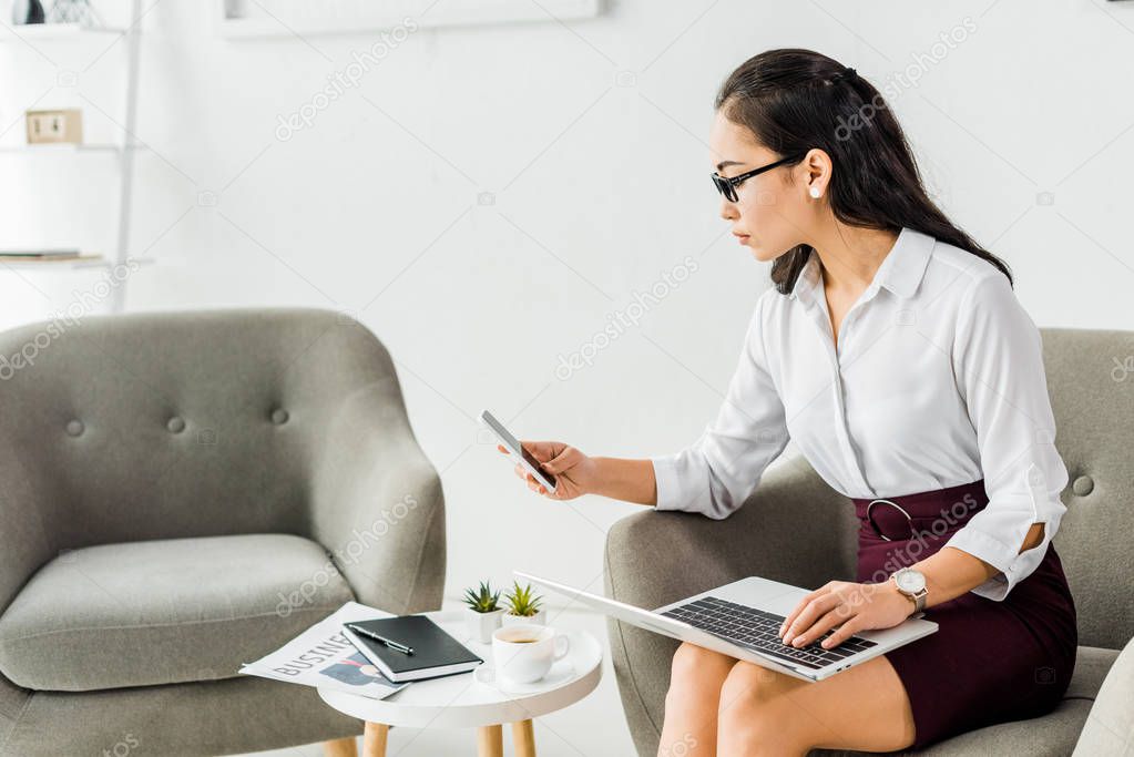  asian businesswoman sitting with laptop and using smartphone in office
