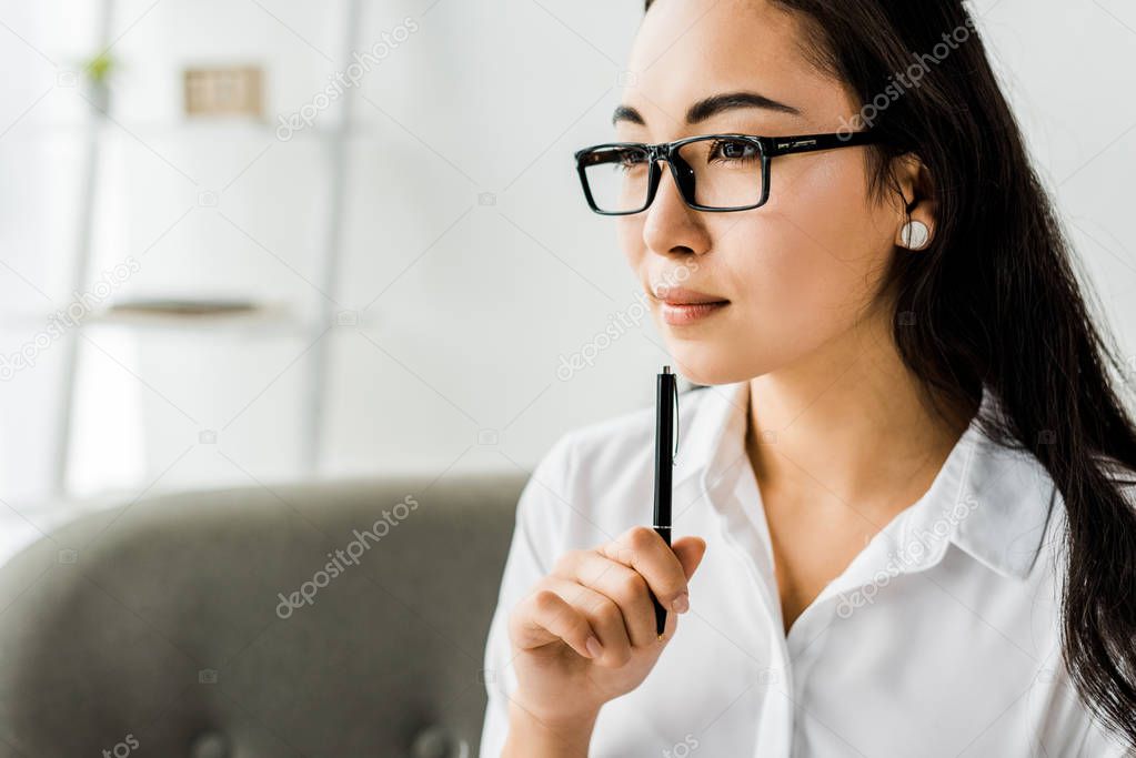 beautiful asian businesswoman in formal wear and glasses holding pen and looking away in office