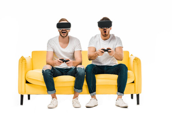 эмоциональные молодые люди в наушниках виртуальной реальности, играющие с джойстиками, изолированными на белом
 