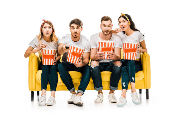 young friends holding popcorn boxes and watching tv while sitting on yellow sofa isolated on white  