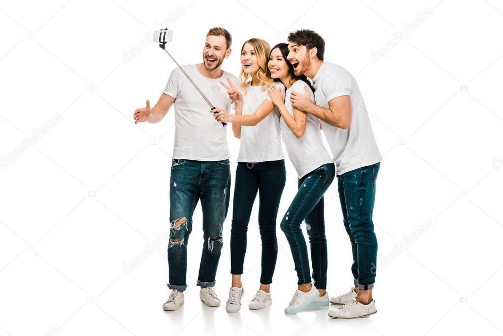 happy young male and female friends standing together and taking selfie with smartphone and selfie stick isolated on white