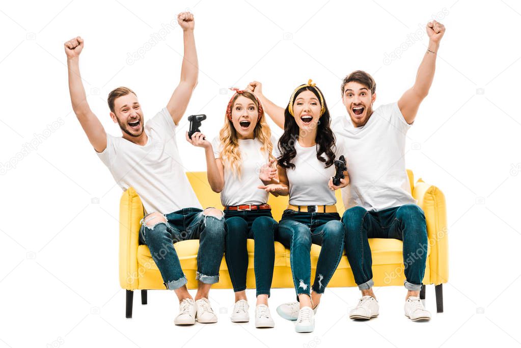 excited young friends sitting on couch and playing with joysticks isolated on white