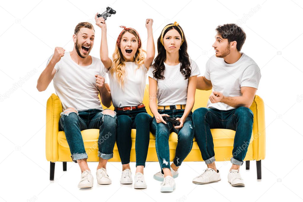 emotional young friends sitting on couch and playing with joysticks isolated on white 