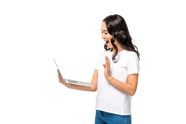 Smiling young asian woman using laptop and showing hello gesture isolated on white