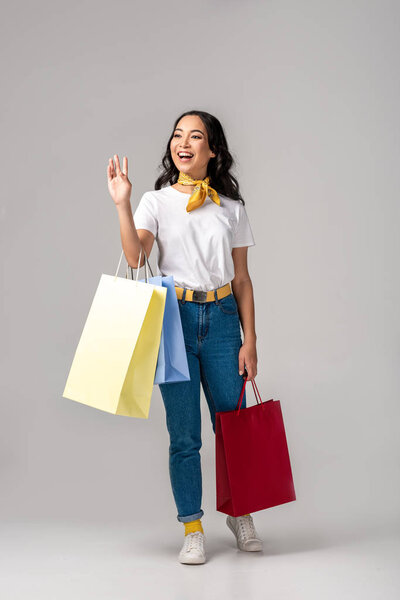 Trendy dressed young asian woman holding colorful shopping bags and waving by raised hand on grey
