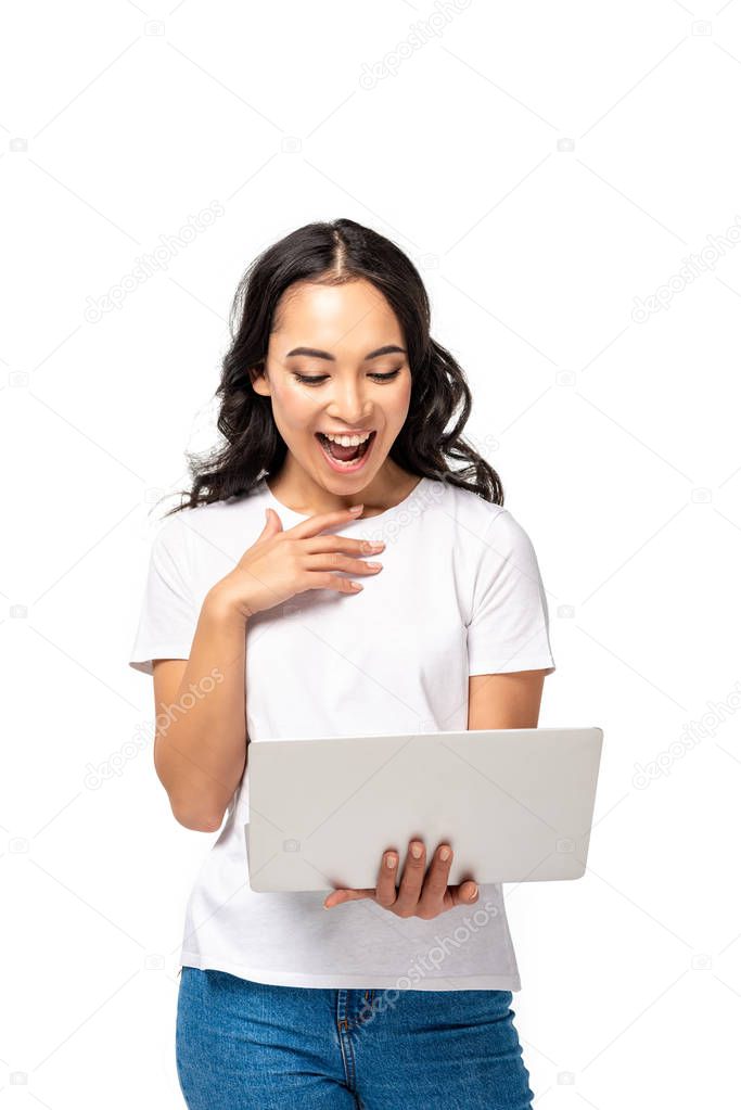 Laughing young asian woman using laptop and holding hand on chest isolated on white