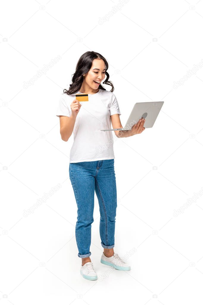 Happy asian woman in white t-shirt and blue jeans using laptop and holding credit card isolated on white