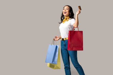 Gülümseyen trendy renkli alışveriş çantaları taşırken ve kredi kartı gri izole tutan Asyalı kadın giyinmiş