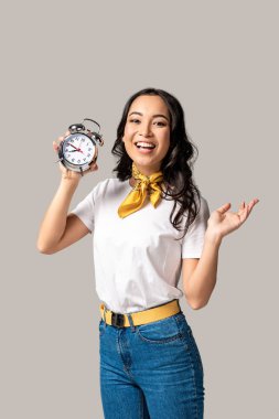 smiling young asian woman in white t-shirt and blue jeans holding alarm clock isolated on grey clipart