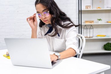 Thoughtful asian freelancer using laptop at home workplace and correcting glasses clipart