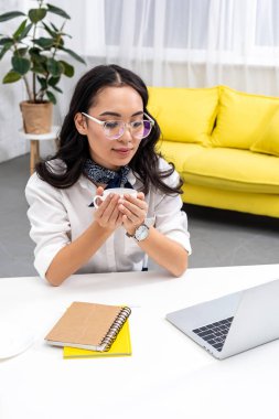 Çalışma masası oturma ve kahve tutan Asya freelancer gülümseyen