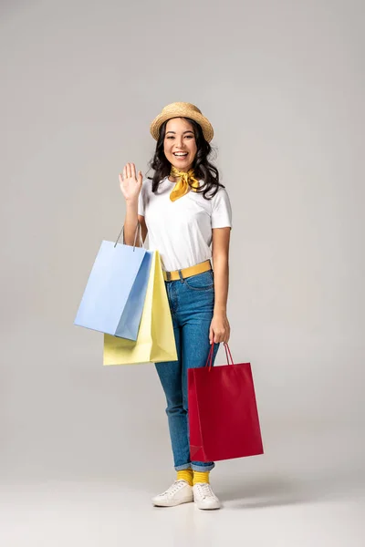 おしゃれな帽子カラフルなショッピング バッグを押しながらグレーに手を振って笑顔幸せなアジア女 — ストック写真