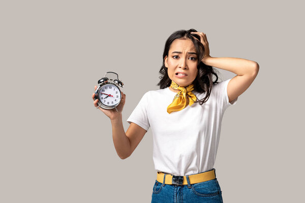 Shocked young asian woman showing alarm clock and holding hand on head isolated in grey 