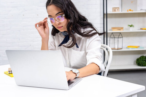 Thoughtful asian freelancer using laptop at home workplace and correcting glasses