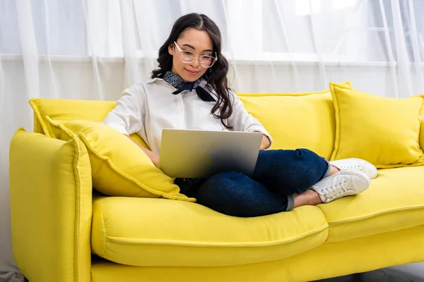 自宅に黄色のソファに足で座りながらノート パソコンを使用してはかなり笑みを浮かべてアジア フリーランサー — ストック写真