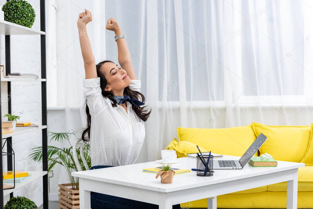 Tired asian freelancer stretching on chair with raised hands while sitting at work desk at home