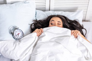 Young beautiful asian woman looking at alarm clock from under the blanket with squinting one eye clipart