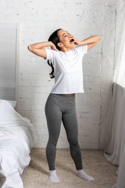Young asian woman in white t-shirt and grey leggings standing in bedroom in yawning clipart