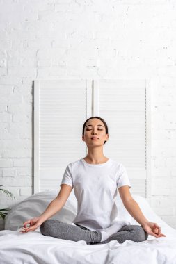Asyalı kadın yatakta lotus poz oturan beyaz tişört ve gri tozluk