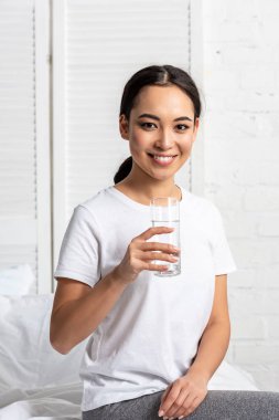 Asyalı kız yatakta bardak su ile oturan beyaz t-shirt gülümseyen