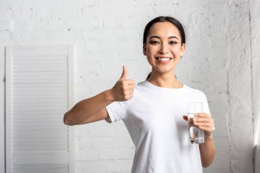 bardak su tutarak ve başparmak gösterilmesini beyaz t-shirt gülümseyen Genç Asyalı kadın