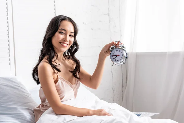 Sorrindo Bela Asiática Mulher Segurando Despertador Enquanto Sentado Cama Olhando — Fotografia de Stock