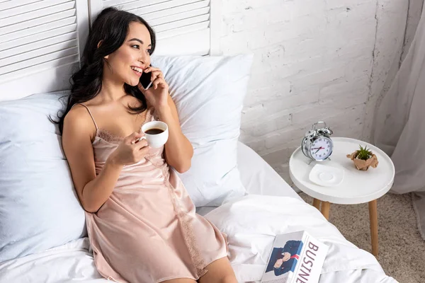 スマート フォンで話していると ベッドに滞在しながらコーヒーを飲む優雅なネグリジェで美しいアジア女性 — ストック写真