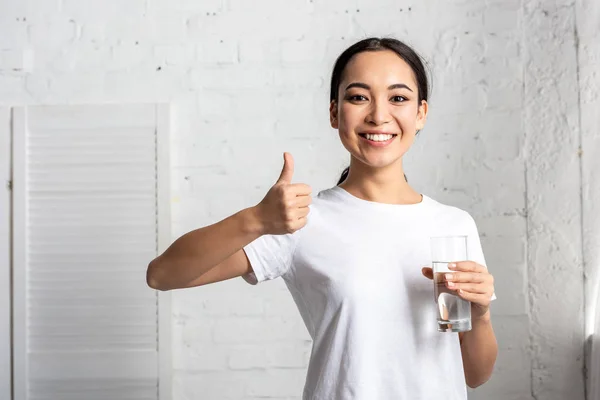 微笑的年轻亚洲女人在白色 T恤举行玻璃的水 并显示拇指 — 图库照片