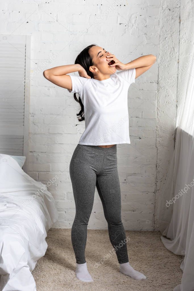 Young asian woman in white t-shirt and grey leggings standing in bedroom in yawning