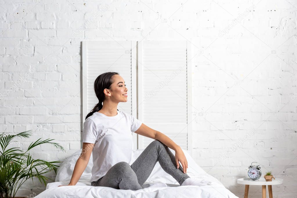 asian woman in white t-shirt and grey leggings sitting in bed and resting after morning exercising