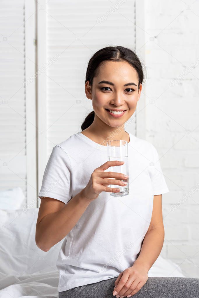 smiling asian girl in white t-shirt sitting on bed with glass of water