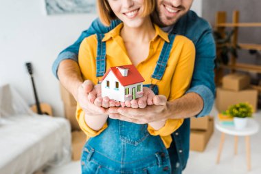 Cropped view of smiling couple holding toy house and looking at camera at home clipart