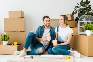 Karton kutuların yanında yerde otururken kahve içen neşeli çift