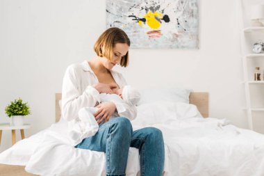 Yatakta oturan ve bebeği emziren kot pantolonla anne 